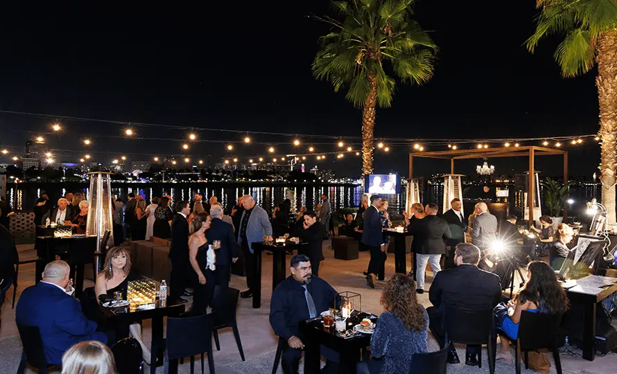 Outdoor event with people gathered around tables, illuminated by string lights and patio heaters.