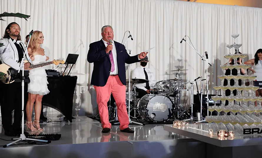 Man speaking into a microphone on stage with a band performing behind him.