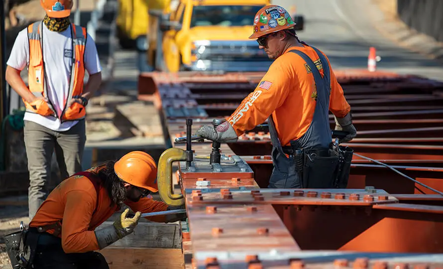 Construction workers assembling steel bridge beams with a large clamp.
