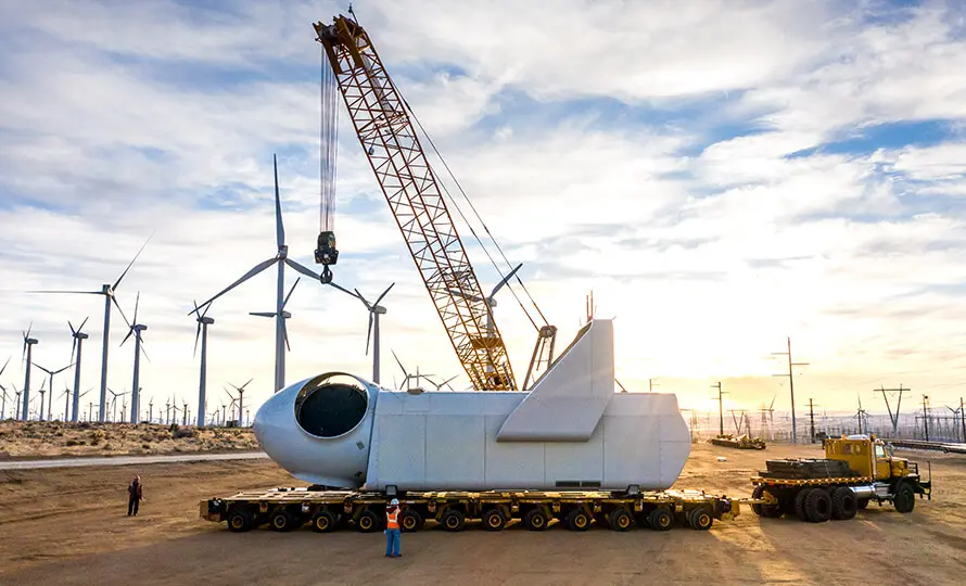 Wind turbine nacelle being transported on a trailer with a crane in the background at a wind farm.