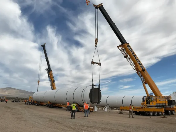 A large crane hoists a substantial pipe onto a truck, showcasing heavy machinery in action at a construction site.