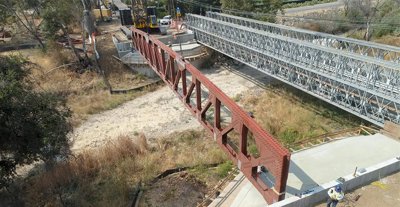 Construction workers securing a steel bridge beam over a natural landscape.
