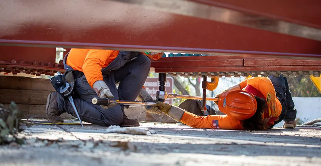 Construction workers tightening bolts under a steel bridge beam.