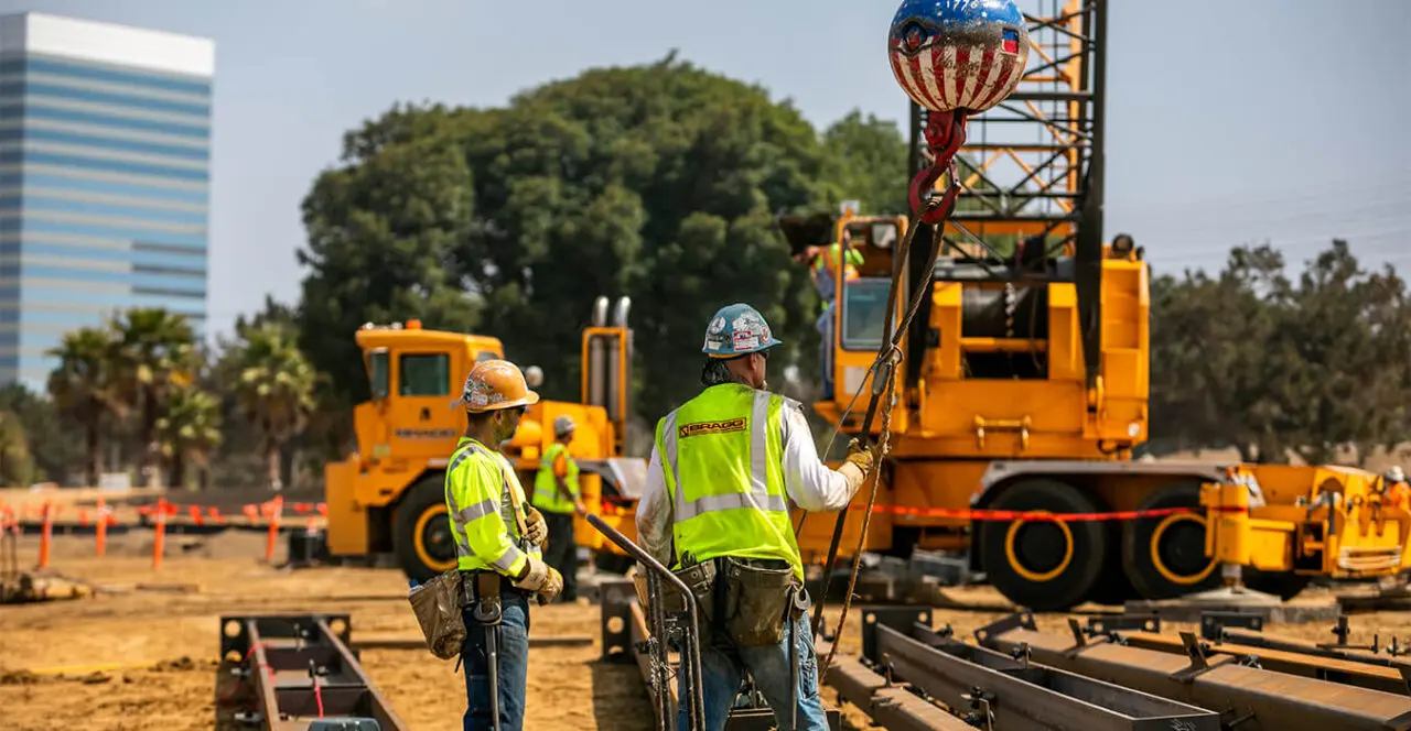 Construction workers aligning steel beams on a construction site with a large crane in the background.