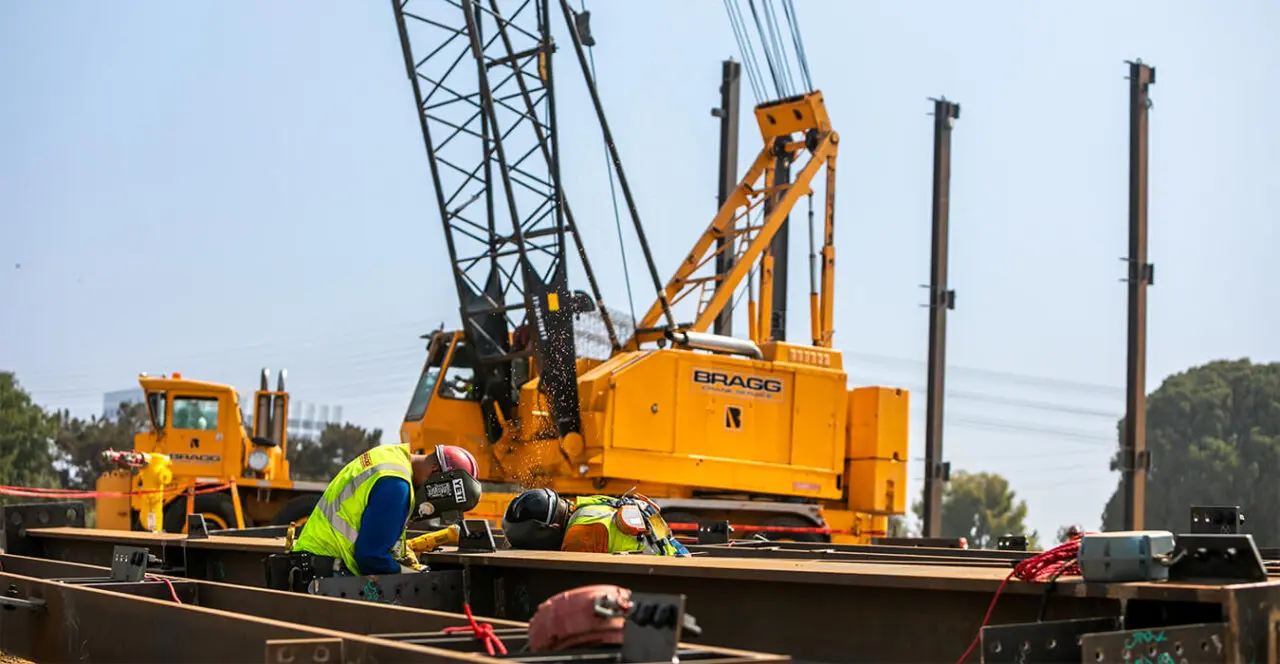 Construction workers welding steel beams with a large crane in the background on a construction site.