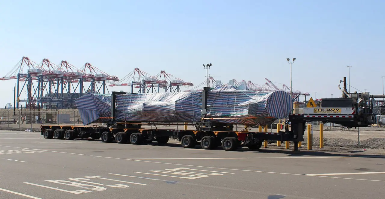 Long flatbed trailer transporting a large covered load through a port area with container cranes in the background.