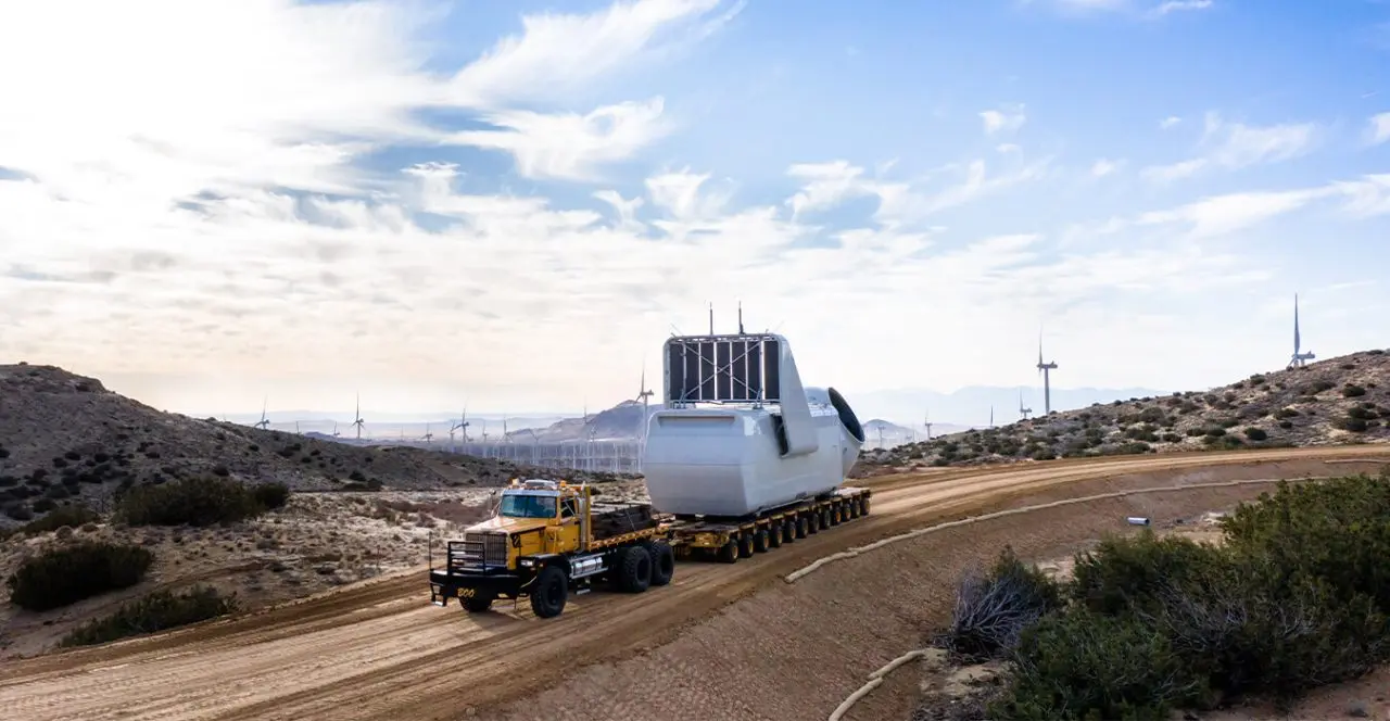 Wind turbine blade transport on a multi-axle trailer through a desert landscape.